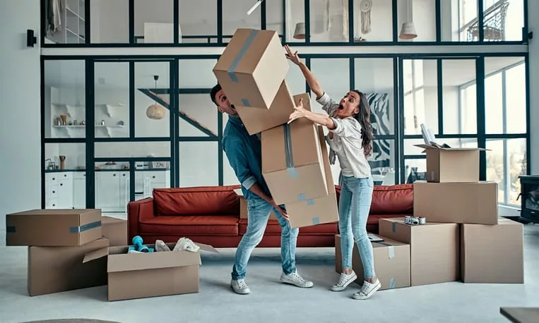 Una coppia che cerca di bilanciare una pila di scatole da trasloco in un ampio soggiorno, simboleggiando la necessità di una soluzione efficiente per un'unità di stoccaggio.